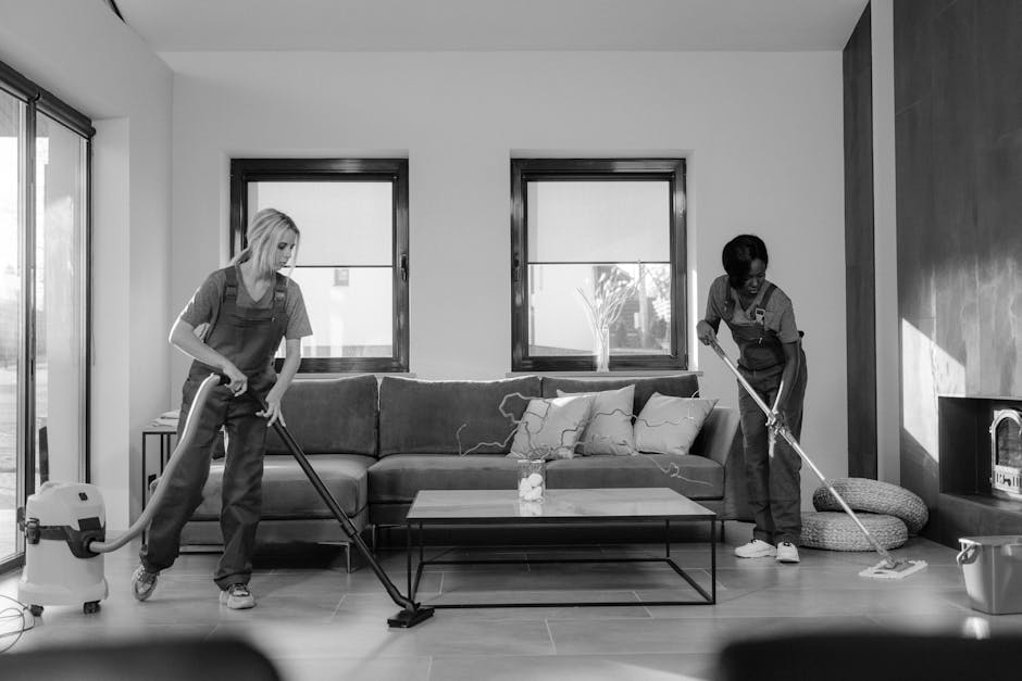 A woman in red overalls is engaged in surface cleaning in a modern kitchen, using a mop on the light-colored tiled floor. The kitchen features sleek white cabinetry with a built-in oven, a black count